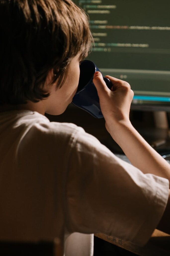 A young coder focused on programming at a computer while sipping coffee.