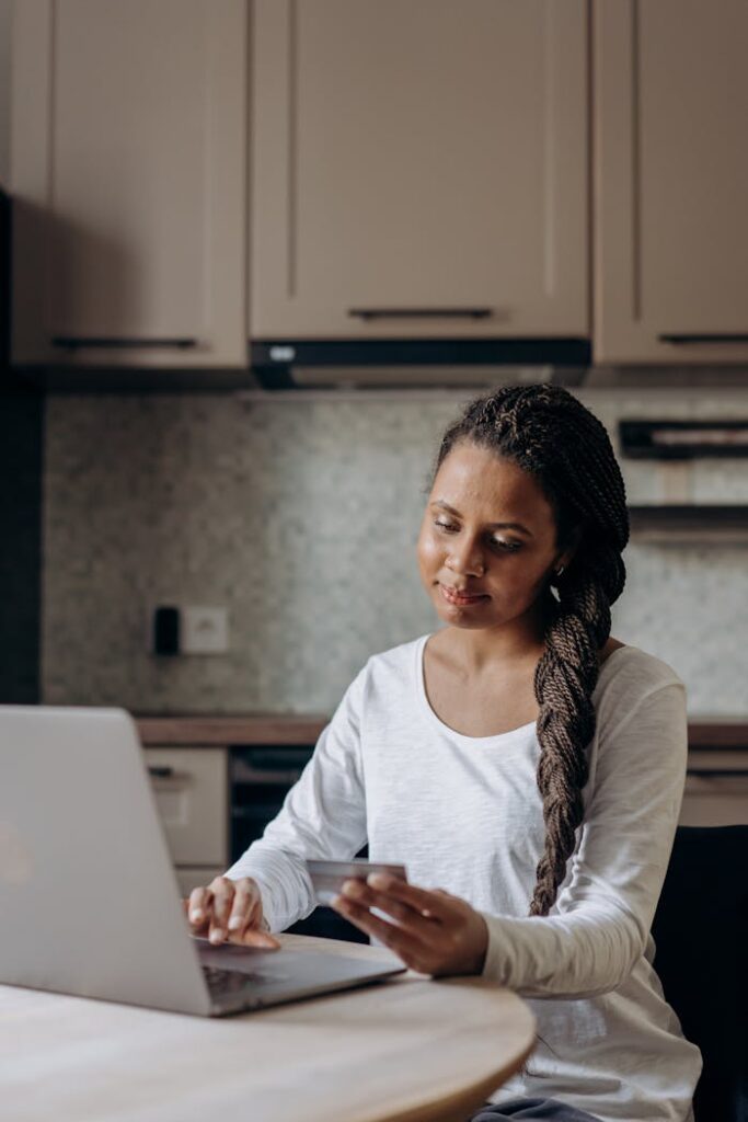 Woman with dreadlocks shopping online using a laptop in her kitchen.
